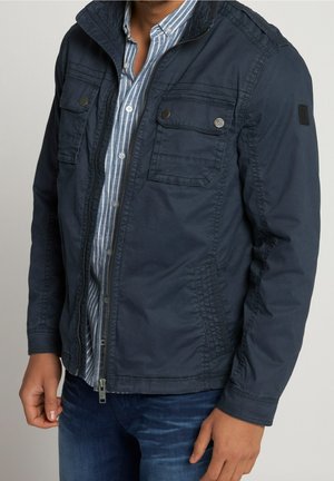 Uomo che indossa una giacca blu navy aperta sopra una camicia a righe blu e bianche con bottoni e jeans blu, in piedi davanti a uno sfondo neutro.