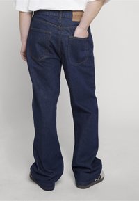 Osoba nosící tmavě modré džínové kalhoty s volnějším střihem, jednou rukou v zadní kapse, v kombinaci s bílou košilí a teniskami.