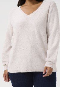 Lyse beige V-hals sweater med en blød tekstur, prydet med spredte sølvfarvede pailletter og ribbede manchetter, båret over mørk denim.