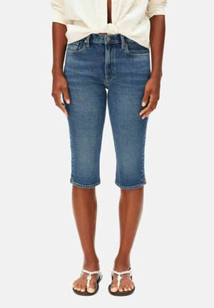 Femme portant un short en denim bleu au genou, une chemise blanche nouée et des sandales blanches tressées, debout devant un fond uni.