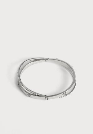 Sølv bangle-armbånd med overlappende bånd, rhinsten og små runde dekorative detaljer, vist på en ensfarvet hvid baggrund.