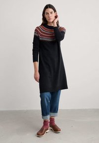 Černé pletené šaty s vzorovanou lérou v oranžové a bílé. Spárováno s modrými ohrnutými džíny a hnědými dřeváky. Měkká textura.
