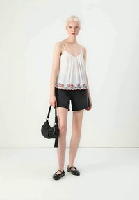 Top branco sem mangas com bordado floral colorido na bainha, combinado com calções pretos e sapatilhas pretas, segurando uma pequena mala preta.
