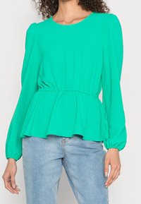 Maglietta verde a maniche lunghe con scollo rotondo, vita elastica e orlo svasato. Tessuto morbido con una consistenza liscia. Abbinata a jeans azzurri chiari.