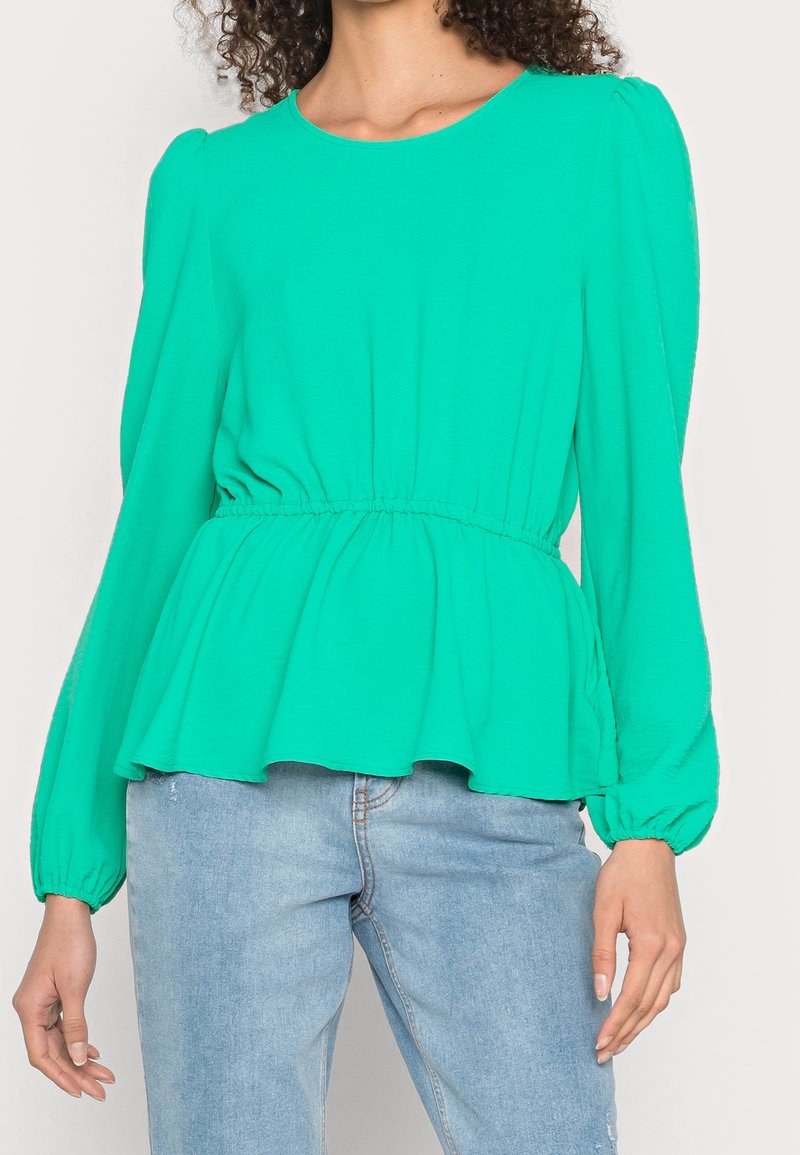 Maglietta verde a maniche lunghe con scollo rotondo, vita elastica e orlo svasato. Tessuto morbido con una consistenza liscia. Abbinata a jeans azzurri chiari.