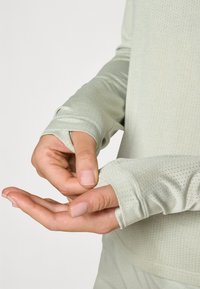 Hosszú ujjú világoszöld ing, textúrált anyagból készült, kis perforációkkal, az ujjak végén hüvelykujjtartókkal.