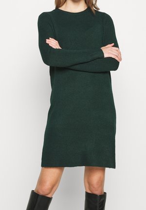 Kobieta w ciemnozielonej dzianinowej sukience z długim rękawem i okrągłym dekoltem oraz czarnych kozakach sięgających kolan, ze skrzyżowanymi ramionami na piersi.