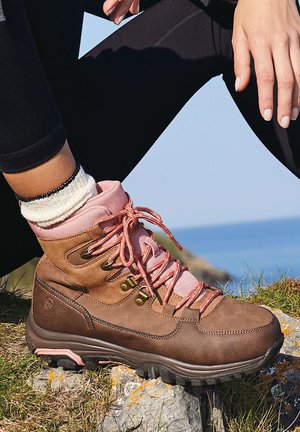 Bota de senderismo marrón con acentos rosas, material de ante y textil, suela resistente y cordones rosas, llevada con calcetines blancos sobre una superficie rocosa.