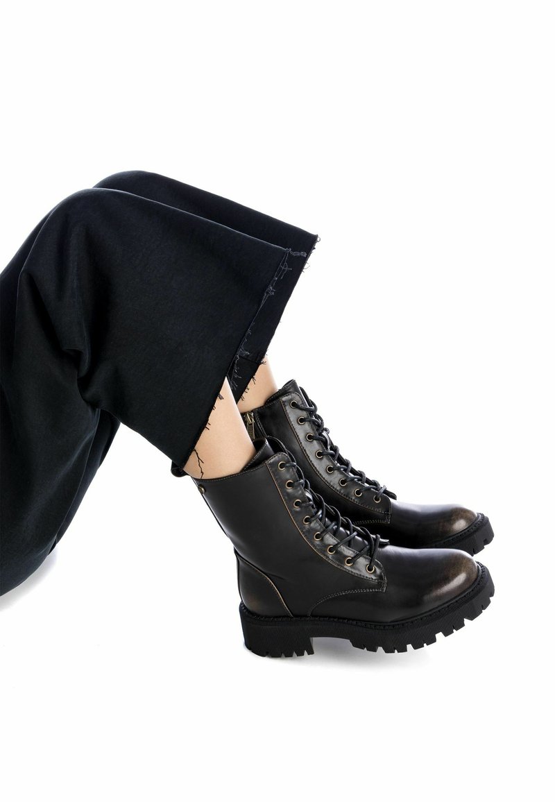 Stivali da combattimento neri in pelle con suola spessa e occhielli metallici, dotati di un design con lacci e una superficie liscia. I pantaloni hanno orli sfilacciati.