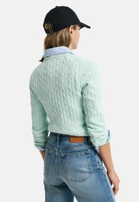 Maglione a trecce verde menta con maniche lunghe, indossato sopra una camicia colletto azzurro chiaro, abbinato a jeans azzurri chiari a vita alta.