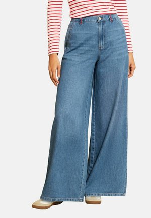 Femeie purtând blugi albaștri cu talie înaltă și pantaloni evazați, bluză cu mâneci lungi în dungi roșii și albe, și adidași albi cu talpă maro.