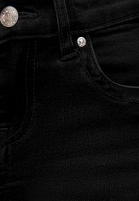 Gros plan sur un jean en denim noir montrant un bouton argenté, une boucle de ceinture et un rivet près de la poche avant.