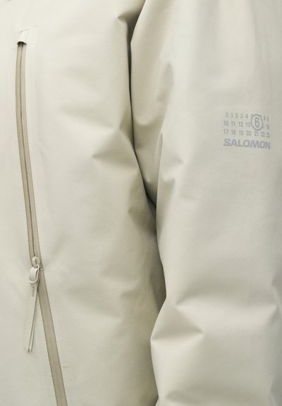 Világos bézs színű, vízálló dzseki sima textúrával, cipzáras zsebbel, diszkrét márkajelzéssel és minimalista dizájn elemekkel.