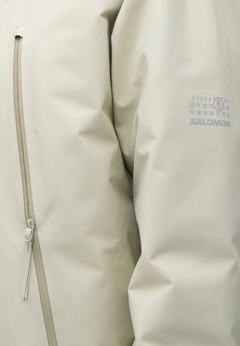 Helle beige, wasserdichte Jacke mit glatter Textur, ausgestattet mit einer Reißverschlusstasche, dezentem Branding und minimalistischen Designelementen.