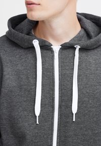 Sudadera gris con una cremallera y cordones blancos. Presenta una textura suave y una capucha amplia. Detalles notables incluyen costuras en contraste.