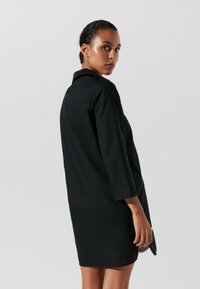 Abito nero con maniche lunghe, colletto e fessure laterali. Texture liscia, design lungo fino al ginocchio, mostrato di spalle con uno sfondo neutro.