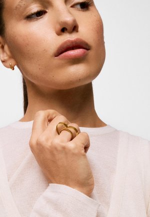 Mujer con piel clara y pecas lleva dos grandes anillos de oro en los dedos, sosteniendo su mano cerca del mentón, vestida con un suéter rosa claro.