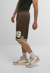 Shorts de sport marron avec une bordure blanche, présentant le numéro 89 en crème et un logo rouge. Le modèle les associe avec des baskets beiges et crème et des chaussettes blanches.