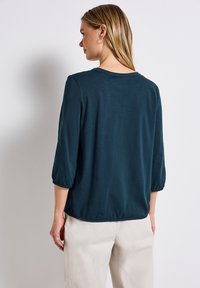 Dunkelblau-teal, leichtes Blusenshirt mit Rundhalsausschnitt, Dreiviertelärmeln mit elastischen Bündchen und lockerer Passform, kombiniert mit hellen Hosen.