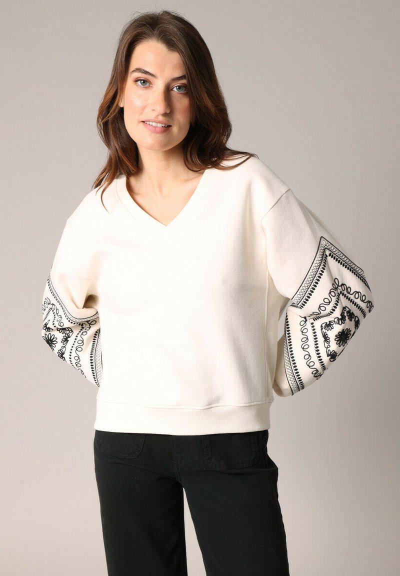 Sweat-shirt crème à col en V avec des manches larges, orné de motifs floraux et géométriques noirs, en tissu texturé et avec un ourlet ajusté.