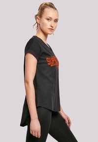 Sort T-shirt med korte ærmer og en let forlænget ryg, der har et orange trykt logo på forsiden. Sammen med sorte leggings.