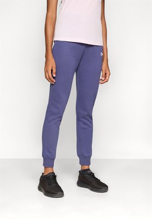 Joggings violets avec une coupe ajustée, fabriqués en tissu doux. Caractéristiques : ourlets élastiques et petit logo sur la cuisse. Associés à des baskets noires.