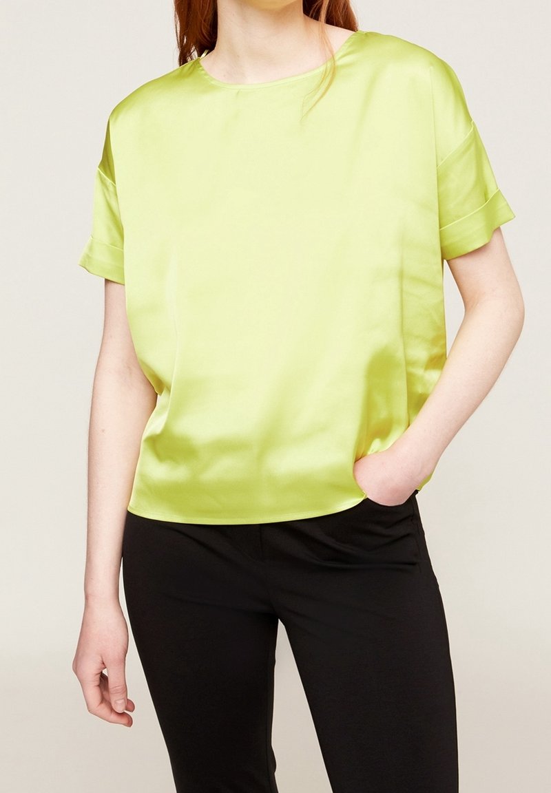 Limetkově zelený saténový top s krátkým rukávem, oblečený kolem krku a volně střižený, s boční kapsou a hladkým povrchem. Kombinovaný s černými kalhotami.
