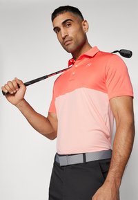 Vīriešu golf polo krekls ar gradienta dizainu korallā un rozā krāsā, izgatavots no viegla auduma. Iekļauj apkakli un logo akcentu. Valkāts ar melniem biksēm.