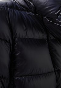 Svart pufferjacka med quiltade sektioner. Glansig, slät yta och metallbeslag för stängning. Har en hög krage-design.