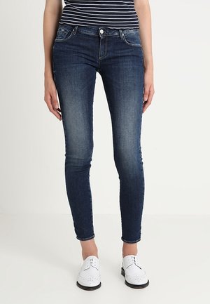 Femme portant un jean skinny bleu foncé, une chemise rayée noire et blanche, et des chaussures blanches à lacets, debout sur un fond blanc.