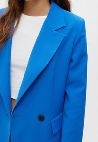 Klart blå blazer i mjukt tyg, med slagkrage, en svart knapp och sidfickor. Bärs över en vit kort topp.