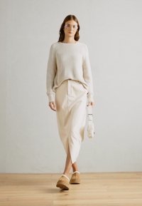 Jersey de rayas en gris claro, falda midi color crema, zuecos marrones, gafas transparentes, sosteniendo un bolso blanco. Suelo de madera y fondo sencillo.
