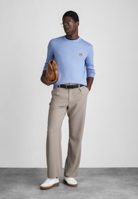 Svijetloplava dukserica s malim logotipom lisice, bež hlače, bijele tenisice, drži smeđu torbu, nosi naočale, stoji na teksturiranom podu.