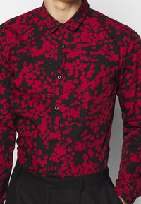 Zwarte shirt met een rood abstract bloempatroon, met knoopsluiting aan de voorkant, klassieke kraag, lange mouwen en een getailleerde pasvorm. Textuurdetails van de stof zijn zichtbaar.