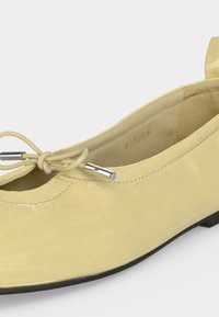 Gros plan d'une ballerine en cuir jaune pâle avec un nœud et un laçage à embout argenté sur fond blanc.