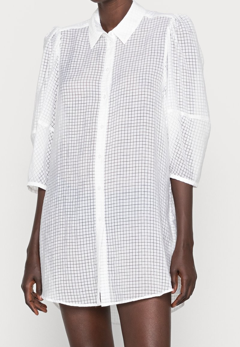 Femme portant une robe chemise blanche à manches longues, boutonnée, avec un motif à grille noire et un col pointu.