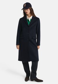 Woman wearing a dark navy long coat, green and white shirt, black jeans, brown boots, and a black cap with a large orange "G" on it.