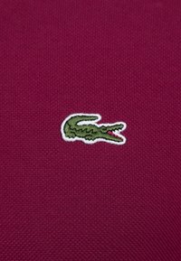 Textil magenta con un patrón texturizado, con un logotipo de cocodrilo bordado en verde y contornado en blanco en el centro.