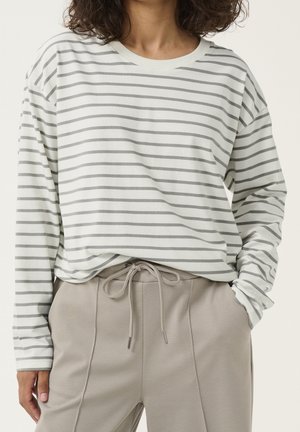 Frau trägt ein weißes Langarmshirt mit grauen horizontalen Streifen und beigen Kordelzughosen, Hand in der Tasche.