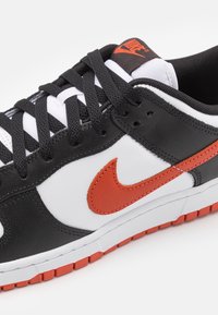 Svart och vit sneaker med en orange Nike swoosh, med en texturerad läderovandel, svarta snörning och en vit mellansula med en orange yttersula.