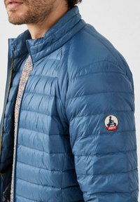 Giacca imbottita blu con design trapuntato, collo alto e toppa rotonda con logo "JOTT" sulla manica sinistra. Materiale liscio e leggero.