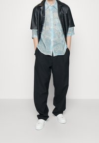 Camisa de cuero negra de manga corta, camiseta de manga larga estampada en azul claro debajo, pantalones anchos negros y zapatillas blancas.
