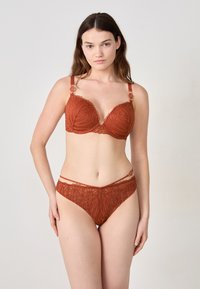 Una modelo posa con un intrincado conjunto de lencería de encaje en color óxido, que presenta un sujetador acolchado con tirantes decorativos y unas braguitas de talle alto a juego.