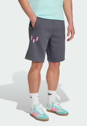 Person trägt graue Shorts mit pinkem Logo, weiße Adidas-Socken und hellblaue und pinke Adidas-Sneakers und steht mit einer Hand in der Tasche.