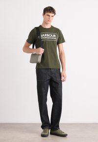 Olivově zelené tričko s bílým logem "Barbour International", spárované s tmavými džíny a olivovými teniskami. Model drží šedou kabelem na rameno.