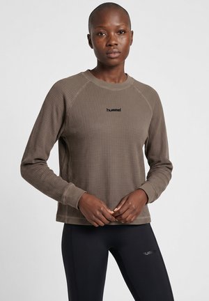 Kvinde med kort hår iført brun tekstureret langærmet trøje og sorte leggings, begge med små "hummel"-logoer, stående mod hvid baggrund.