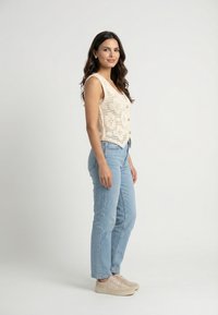 Mujer con cabello largo y oscuro, vestida con una camiseta sin mangas de crochet beige, jeans azul claro y zapatillas beige, de pie contra un fondo liso.