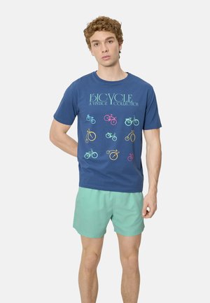 Joven con camiseta azul con ilustraciones vintage coloridas de bicicletas y pantalones cortos verde claro, de pie contra un fondo liso.