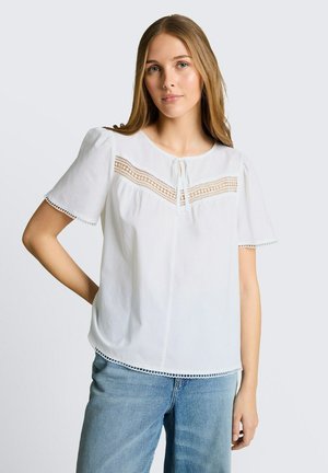 Jeune femme portant un chemisier blanc à manches courtes avec un empiècement en dentelle et un jean bleu, debout avec une main sur la hanche devant un fond uni.