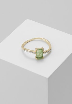 Bague en or avec une pierre précieuse verte rectangulaire au centre et de petites pierres claires incrustées le long de l'anneau, sur un bloc d'exposition blanc.